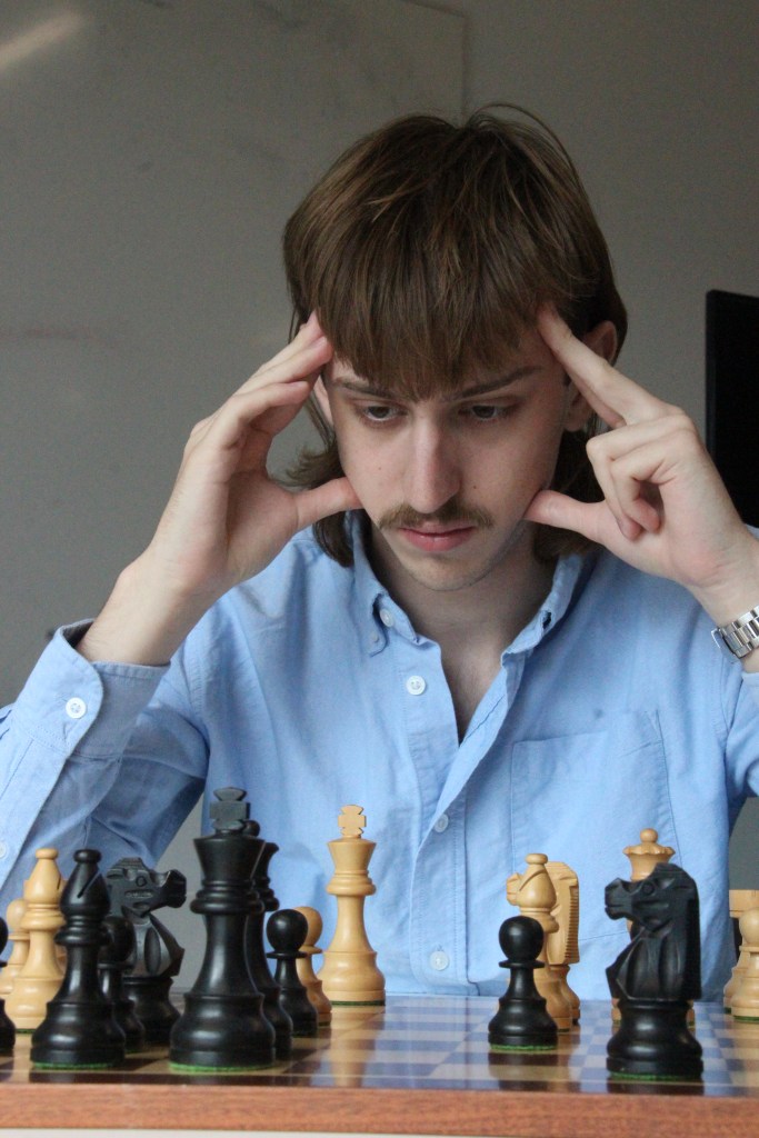 A focused individual chess player in a light blue shirt heavily engaged in chess with beige and black chess pieces.