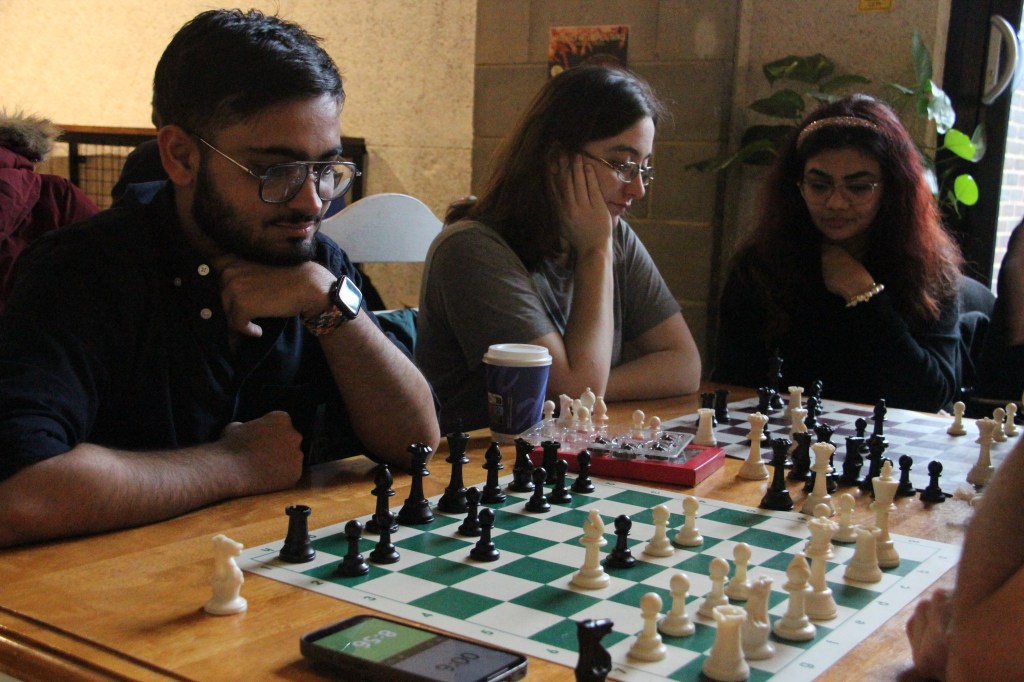 A group of young adults seated at a table, deeply focused on a chess game. A man wearing glasses and a smartwatch ponders his move while two women sit next to him, immersed in thought. A green-and-white chessboard with pieces in play is in the foreground.