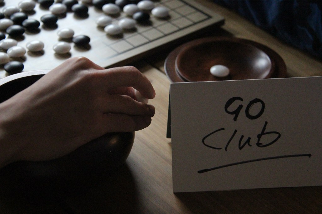 A match of Go in the corner with a sign reading 'Go Club' and a hand lingering over one of the bowls with captured pieces inside.
