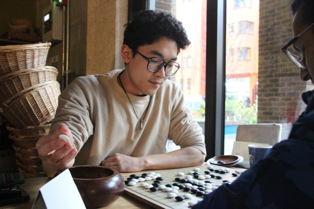 An individual with a cream jumper playing Go against another player, deep in thought.