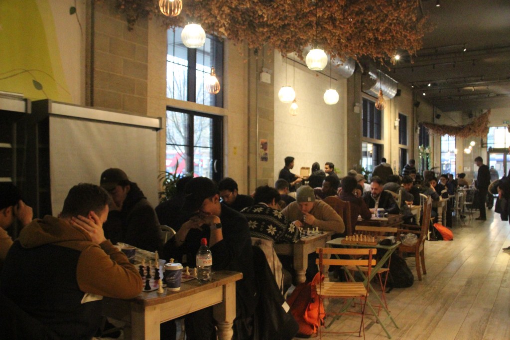 A lively café filled with people engaged in a chess tournament, with wooden tables and chairs, warm pendant lighting, and a relaxed yet focused atmosphere. The room is bustling with players deep in thought as they concentrate on their games.